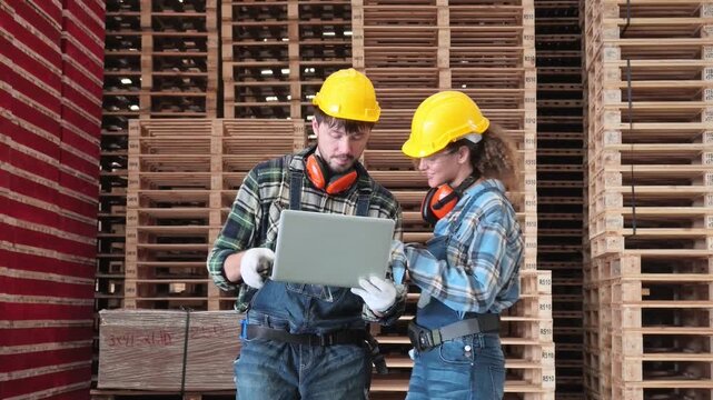 Two factory workers wearing safety helmets and hearing protection review information on a laptop among stacked wooden pallets in a timber warehouse.
