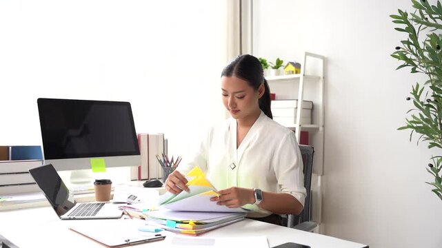 Focused Workstation: A diligent woman in professional attire sits at her desk, engrossed in organizing documents and papers.