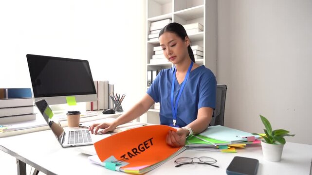 Focused Professional at Work: A young woman in scrubs intently focuses on her laptop at her desk, a folder labeled TARGET lying open in front of her, amidst a backdrop of office supplies and shelves.