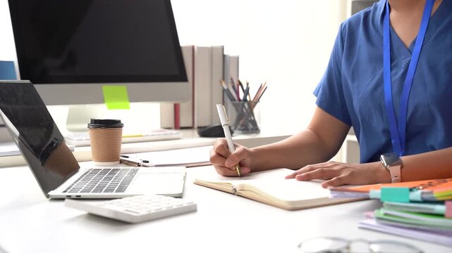 Medical Professional at Desk: A dedicated medical professional, wearing scrubs and a lanyard, diligently takes notes in a planner at a cluttered but organized desk with a laptop, computer monitor.