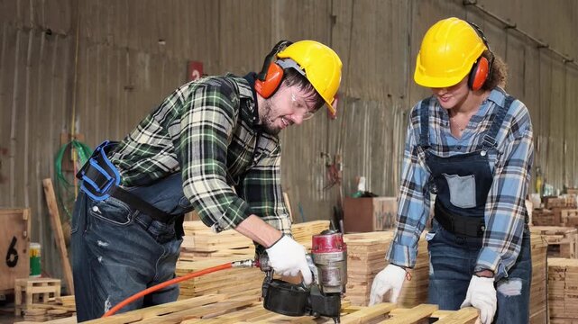 Two carpenters in safety helmets and protective gear review a laptop together inside a lumber factory, surrounded by stacked wood and construction materials.