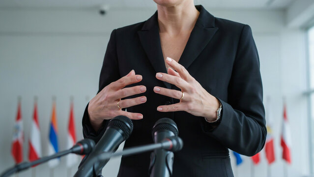 Politician in black suit speaking at press conference with microphones and international flags
