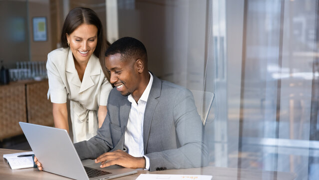 Multiethnic team members working together using laptop in corporate office, mentor helping with project task to apprentice, collaborating, reviewing new business platform or application in workplace