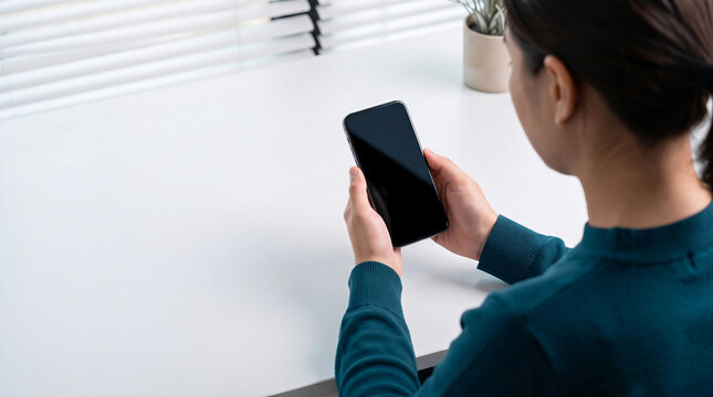 Woman holding a smartphone with a blank screen sitting at a white desk 