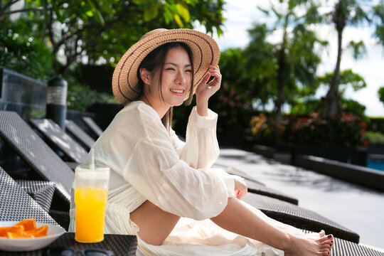 Natural light portrait of relax young asian woman wear swimsuit with hat sitting on a chair by poolside aesthetic style fashion vibes on summertime