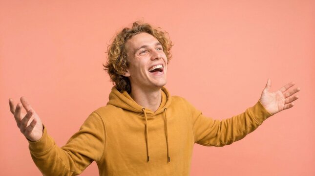 Excited young blonde man in yellow hoodie laughing out loud with arms wide open isolated on bright coral pink studio background portrait