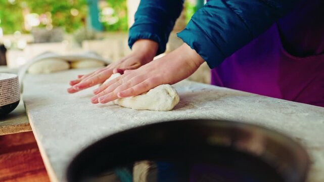 A baker's hands roll a long piece of soft dough on a stone slab beside the dark rim of a traditional Georgian tone oven.