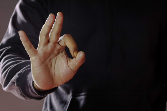 Cropped shot of an Asian man wearing a black sweater, gesturing an OK sign, symbolizing success and approval in a dark setting. Concept of positive non-verbal communication, close-up.