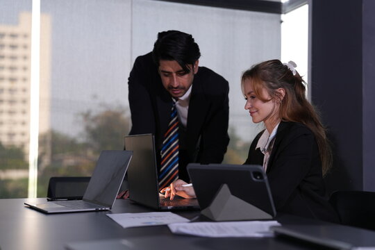 Attentive young businessman bends beside a seated ginger haired woman as they read her tablet screen together at a window side desk in the daylit office in a bright modern coworking office.