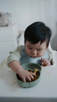 Asian baby boy eating boiled corn on high chair, BLW (baby-led weaning) concept, healthy solid food introducing to infant at home	
