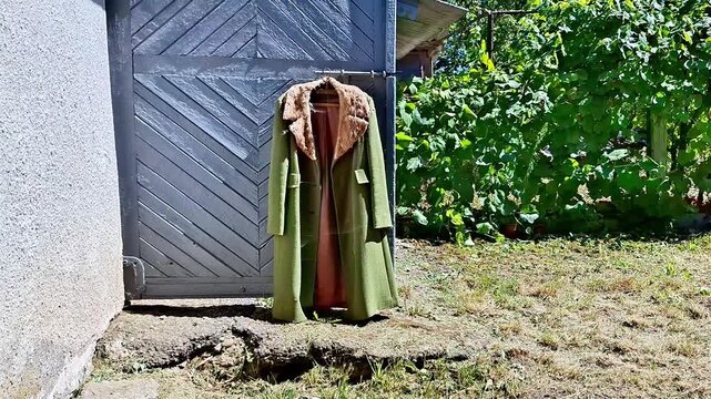 Old green coat hanging on a rustic wooden garden gate beside a house, surrounded by greenery and dry grass. Outdoor rural scene showing an abandoned or unused garment