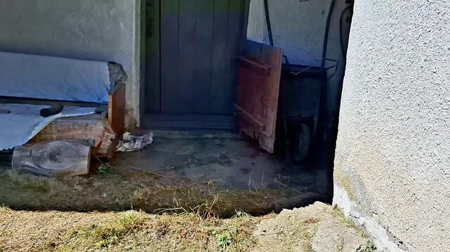 Weathered entrance to an old rural shed with a wooden door, worn walls, scattered household items and dry grass, highlighting an aged countryside property and neglected outdoor space