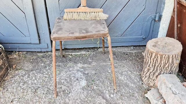 Wooden stool with a broom resting on top, standing beside a tree stump in front of blue wooden doors. A rustic outdoor scene with worn surfaces, simple tools, and traditional surroundings