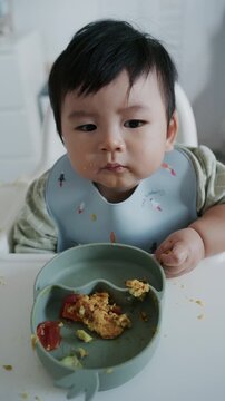 Asian baby boy eating boiled corn on high chair, BLW (baby-led weaning) concept, healthy solid food introducing to infant at home	
