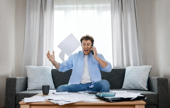 Angry man on phone throws overdue utility bills in despair. He faces severe financial pressure, rising energy expenses, and an economic crisis fueled by inflation.