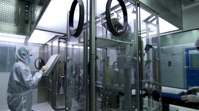 Professional pharmaceutical factory worker and quality control inspector in sterile protective clothing operating automated vaccine production line, automated packaging machinery equipment