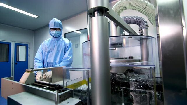 Professional pharmaceutical factory worker quality control inspector in sterile protective clothing operating touchscreen control panel, automated packaging machinery manufacturing facility industrial