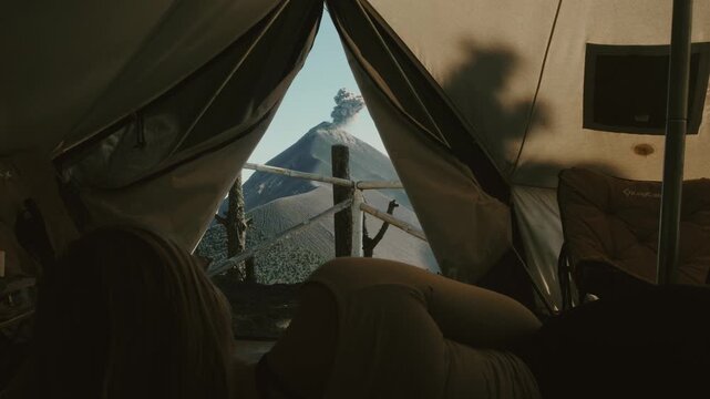 A girl relaxes inside a cozy tent, gazing at Guatemala's erupting Fuego volcano through the tent opening. Morning light, cinematic wide angle, adventure camping on Acatenango trail.