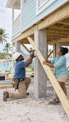Obraz premium Two construction workers install wooden supports beneath an elevated house near a tropical waterfront. The scene shows teamwork and residential construction in progress.