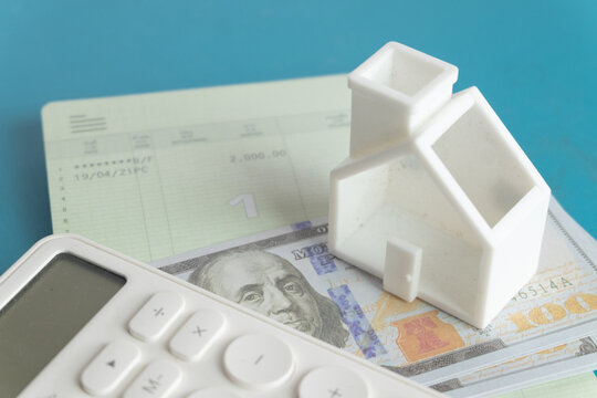 Close up of a white miniature house model, calculator and bank passbook on US dollar bills over a blue background. Concept of real estate investment, calculating property mortgage and saving.