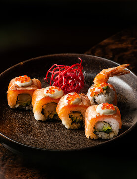 Gourmet salmon sushi rolls topped with creamy sauce, red caviar, and ebi tempura shrimp in a dark ceramic dish.