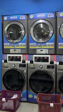 A row of modern washing machines and dryers in a laundromat. The machines are silver with digital displays. Two red laundry baskets are placed in front.