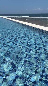A clear blue swimming pool with shimmering water reflects sunlight. The pool is adjacent to a sandy beach and the ocean in the background.