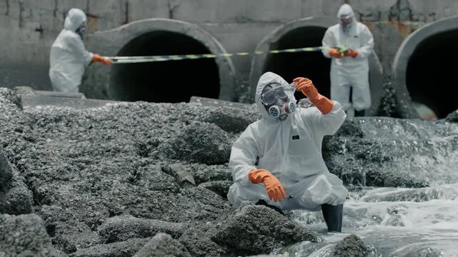 Team in hazmat suits surveys polluted water runoff. One worker kneels, adjusting gear. Others stand at large concrete outflows