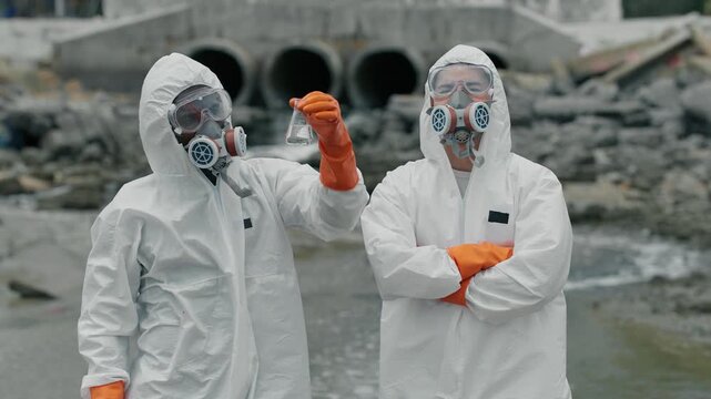 Two scientists in full hazmat gear stand beside polluted water. One holds a vial, examining its contents closely. They wear orange gloves, masks