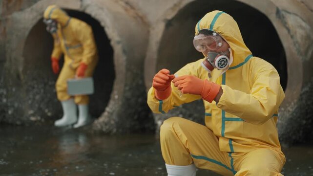 Workers in yellow suits inspect sewage pipes. One crouches, examining something close. Another stands nearby, holding a container. Both wear masks