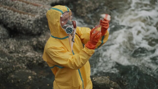 Scientist examines water sample near rocky coast. Waves crash behind the researcher in full gear. Yellow suit shields against environmental hazards