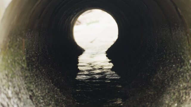 Dark tunnel leads to bright, circular light beyond. Water flows steadily inside concrete pipe. View through wet, rough interior walls