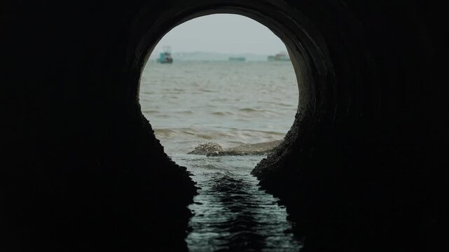 Dark tunnel leads to choppy sea waters. Water flows steadily through circular opening. Distant ships move across grayish horizon