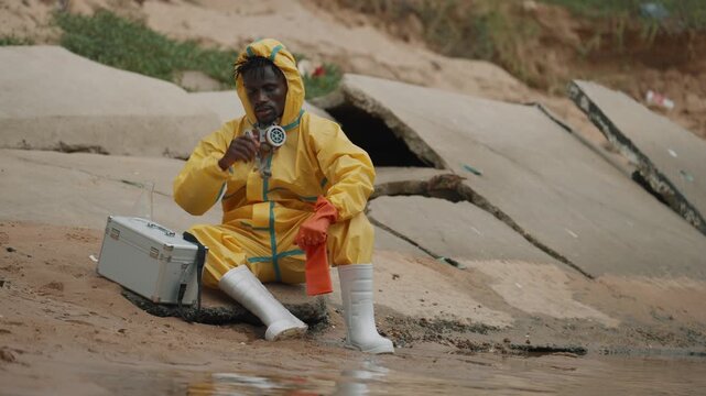 Man in yellow hazmat suit kneels beside muddy water. He wears protective gear and gloves. He adjusts his mask while seated beside a white case