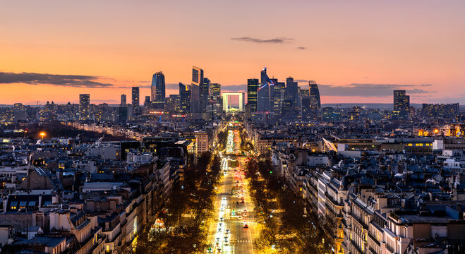 Panoramic view of the La Defense financial district and avenue traffic in Paris, France at sunset with illuminated skyscrapers.