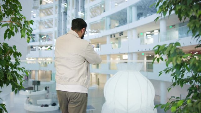 Man seen from behind standing at glass railing in modern multi level atrium holding smartphone with both hands and looking down