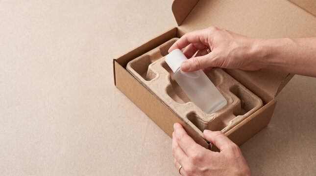Hands removing an unbranded skincare bottle from an open cardboard box with molded pulp protective packaging. Sustainable cosmetic packaging and eco-friendly unboxing concept with copy space.
