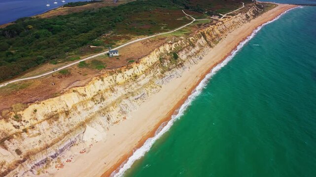 Aerial footage along the cliffs and beach at Hengistbury Head in Dorset, England. The shot shows the exposed coastal cliff face, heathland above, sandy shoreline and turquoise waters of Christchurch B