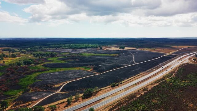 Drone footage of a large, dark burn scar covering heathland and rough grassland, running right up to the edge of a busy dual-carriageway with moving traffic, surrounded by unburnt woodland, green fiel