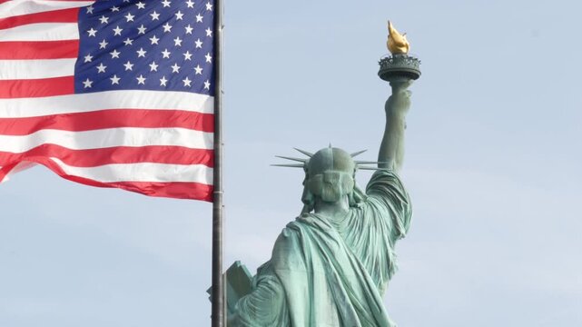 Statue of Liberty on Liberty Island, New York, United States. Cooper monument from behind, democracy and patriotism symbol near Manhattan, NYC USA. American flag waving in wind. Independence concept.