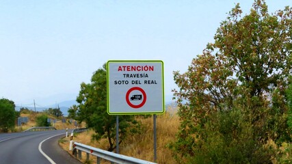 An informational road sign located beside an asphalt road in Spain—specifically regional route M-608—bears the inscription "Atención Travesía Soto del Real,"September 10, 2026  © GenadiyGM