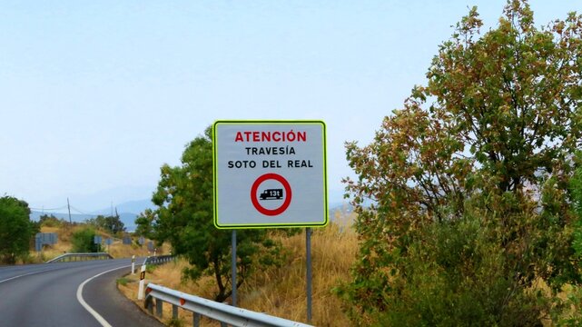 An informational road sign located beside an asphalt road in Spain&mdash;specifically regional route M-608&mdash;bears the inscription "Atenci&oacute;n Traves&iacute;a Soto del Real,"September 10, 2026 