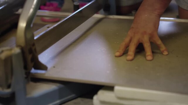 Close-up of a worker holding a large gray floor tile on a manual rail cutter, positioning the slab for accurate scoring and sizing during renovation.
