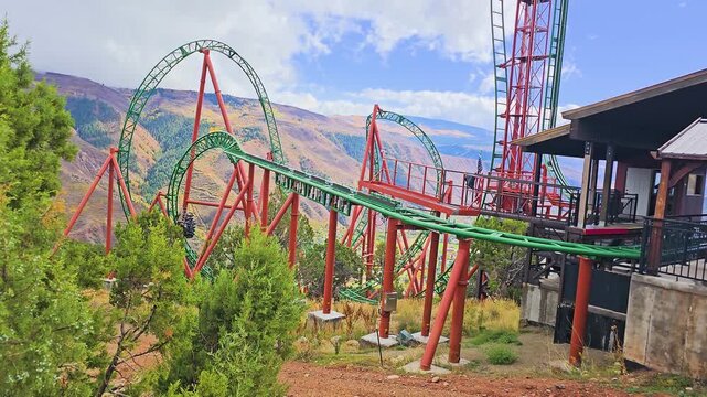 Steel coaster train races through a tight inversion at a mountain amusement park in Colorado, surrounded by looping green track, red columns, pine trees, and steep alpine terrain.