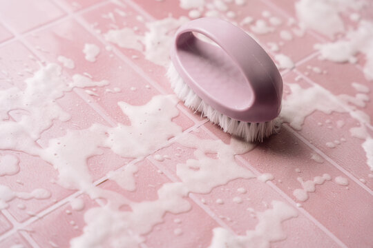 Purple scrubbing brush resting on foamy pink tiles with white bristles.
