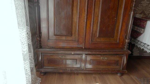 Vintage wooden wardrobe in cozy bedroom interior with lace curtain, patterned wallpaper, warm daylight, worn carved details, paneled doors, lower drawers, and rustic domestic charm