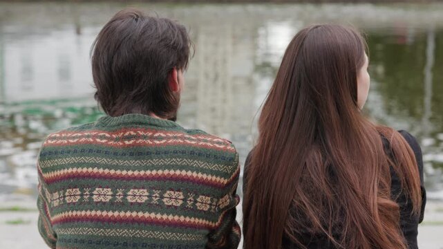 Two people seated on bench by pond with patterned sweater and long brown hair gesturing toward water from behind