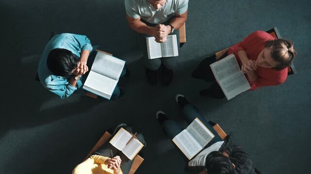 Top view of skilled diverse prayer clasping hand and praying to god while sitting at chair in circle. Top down aerial view of people folded hand while sitting with opened bible on laps. Symposium.