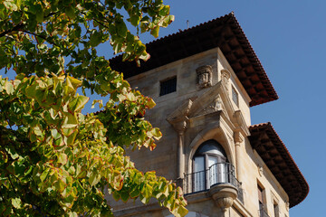 Sunlit corner of a historic stone building in Gijon with a wrought iron balcony, carved crest and tiled eaves behind green tree leaves. Heritage facade detail for travel and tourism marketing. © Renata
