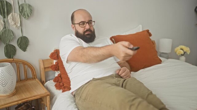 Man, bald with beard, wearing white t shirt, holding remote pointing at tv while reclining on bed in bedroom; relaxation.